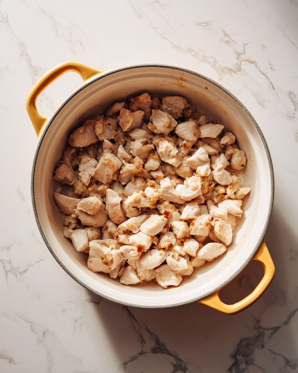 A white pot with a yellow handle filled with small pieces of cooked chicken that are light brown and slightly shiny, spread unevenly across the pot surface with some browned bits stuck at the center. The pot sits on a white marbled surface. photo taken with an iphone --ar 4:5 --v 7