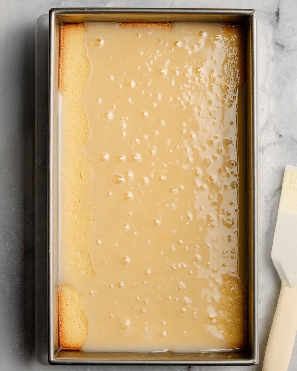 A metal rectangular pan filled with one smooth layer of light golden cake soaked evenly with a shiny, pale cream-colored syrup that covers the entire surface. The cake texture shows small, soft bubbles underneath the thick syrup, and the edges of the cake are slightly darker. The pan sits on a white marbled surface with a white spatula handle partially visible at the bottom right corner. Photo taken with an iphone --ar 4:5 --v 7