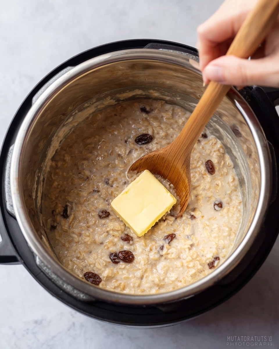 Inside a silver metal pot with a black rim, creamy oatmeal mixed with raisins is being stirred by a wooden spoon. A woman’s hand holds the spoon, which carries a square piece of yellow butter melting on top of the oatmeal. The oatmeal has a thick, slightly textured look with small chunks visible, and some brown specks scattered throughout. The pot sits on a white marbled surface with soft light highlighting the rich, warm colors of the dish. photo taken with an iphone --ar 4:5 --v 7