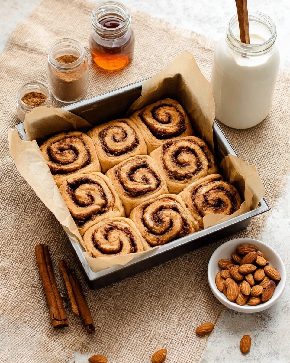 A metal square baking pan lined with light brown parchment paper holds nine golden brown cinnamon rolls arranged in three rows. Each roll has a deep brown spiral swirl of cinnamon filling visible in the soft, fluffy dough. Near the pan, a small white bowl is filled with whole almonds, with some almonds scattered nearby. To the left, there are cinnamon sticks and a nutmeg seed on a white marbled surface. Behind the pan, a glass jar with white icing has a wooden spoon inside, and next to it is a small jar of cinnamon powder and a glass bottle with amber liquid, all set on a light tan textured cloth. photo taken with an iphone --ar 4:5 --v 7
