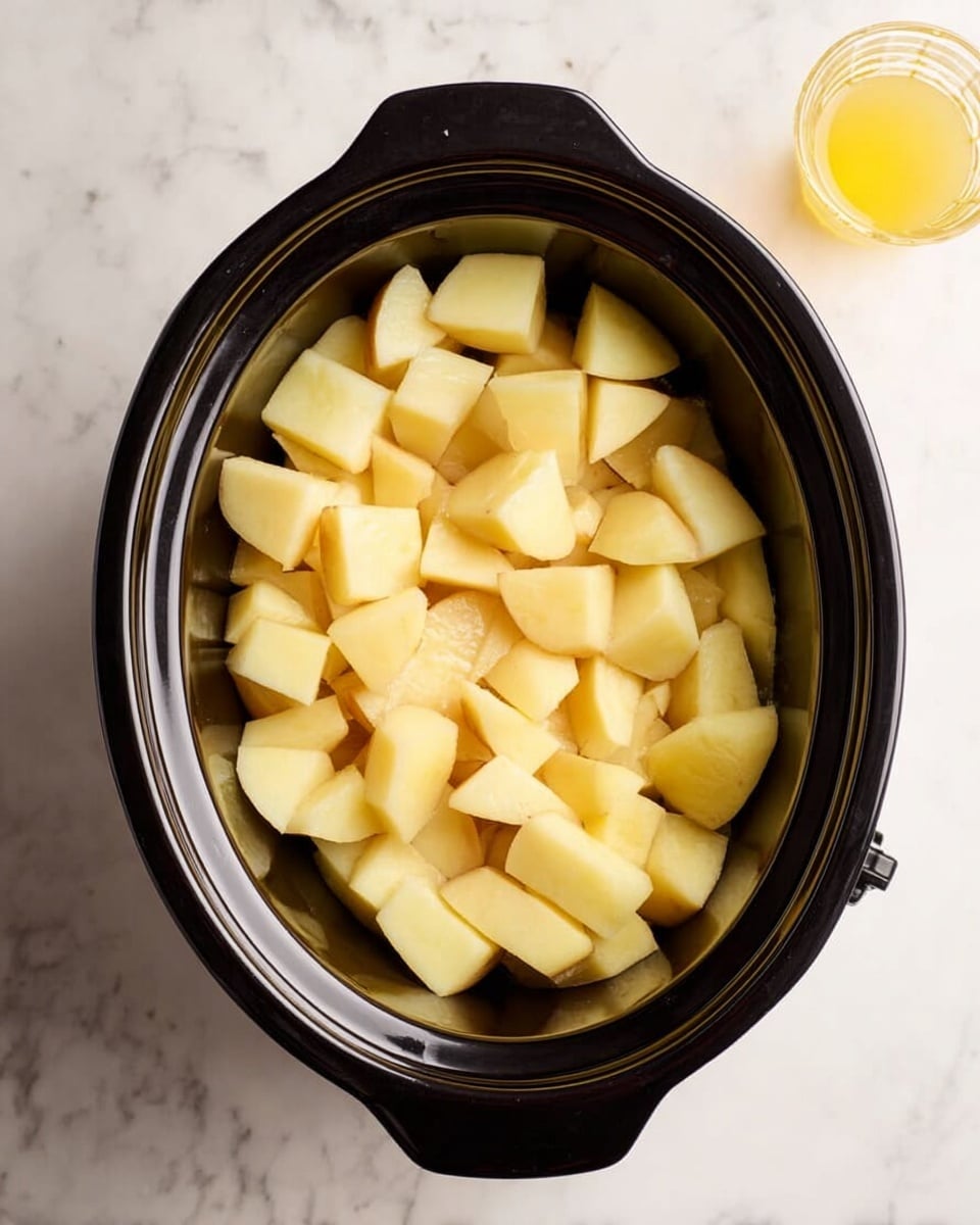 The image shows a black slow cooker filled with one layer of peeled and chopped potatoes, cut into medium chunks with a pale yellow color and smooth texture. The slow cooker is placed on a white marbled surface, with a small glass of yellow liquid on the top right side. The photo is taken from above, capturing the entire slow cooker and part of the surface around it. Photo taken with an iphone --ar 4:5 --v 7