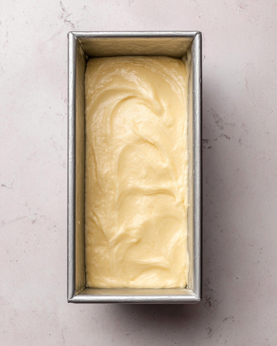 The image shows a rectangular silver metal loaf pan filled with smooth, pale yellow batter. The batter surface is spread evenly with a few soft peaks and subtle swirls, showing a creamy texture. The pan is placed on a white marbled textured surface that contrasts softly with the pale batter. Photo taken with an iphone --ar 4:5 --v 7