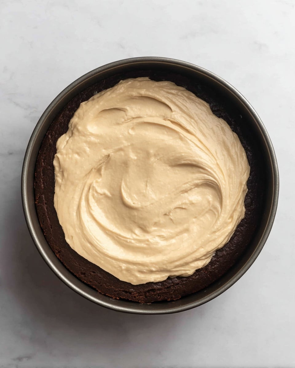A dark brown cake base layer fills the round black baking pan. On top, there is a thick, creamy layer of light tan frosting spread unevenly, showing smooth but slightly whipped texture with a few swirls. The cake and frosting take up the full pan, with frosting covering about half of the cake surface. The pan is placed on a white marbled background. Photo taken with an iphone --ar 4:5 --v 7