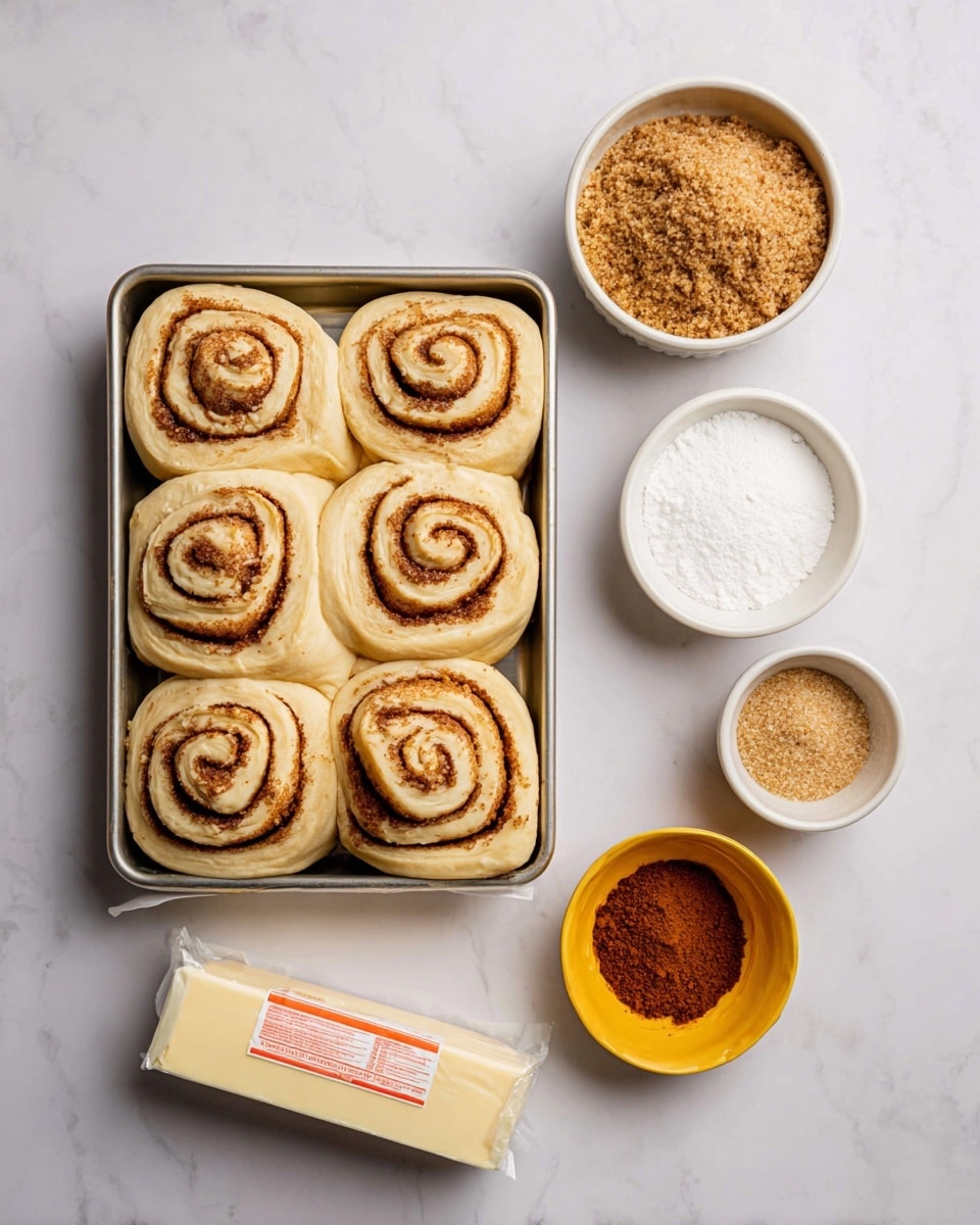 A silver tray filled with eight unbaked cinnamon rolls, each swirl showing light brown cinnamon sugar filling layered within pale dough, sits on a white marbled surface. To the right, three small white bowls hold ingredients: the top bowl contains light brown sugar, the middle bowl white granulated sugar, and below them, a small yellow bowl with a heap of ground cinnamon. Below the tray sits a stick of butter wrapped in white packaging with red text and measurement marks. The scene is clean and bright, with all items arranged neatly. photo taken with an iphone --ar 4:5 --v 7