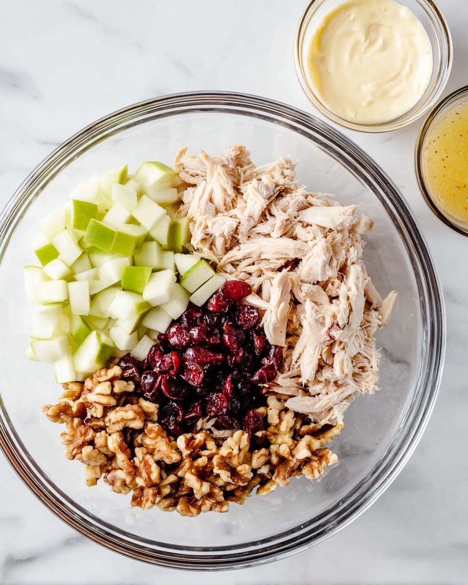 A clear glass bowl with four separate piles of ingredients arranged inside: light brown shredded chicken on the right, small green apple cubes at the top, dark red dried cranberries on the left, and chopped light brown walnuts at the bottom. The bowl sits on a white marbled surface. To the right of the bowl, there are two small glass bowls, one with a light creamy mixture and the other with a yellow substance. Photo taken with an iphone --ar 4:5 --v 7