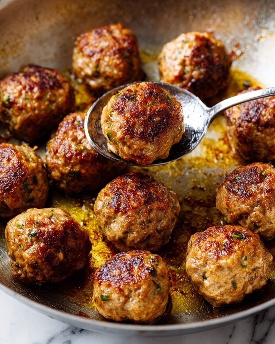 The image shows a close-up of a metal pan filled with 12 golden-brown meatballs. Each meatball has a slightly rough, browned surface with visible small bits of herbs and spices. The meatballs are arranged closely but not touching, with one meatball being lifted by a metal spoon with a shiny surface reflecting the meatball’s texture. The pan surface looks slightly oily with some browned spots and crumbs around the meatballs highlighting the cooking process. The background is a white marbled texture. photo taken with an iphone --ar 4:5 --v 7