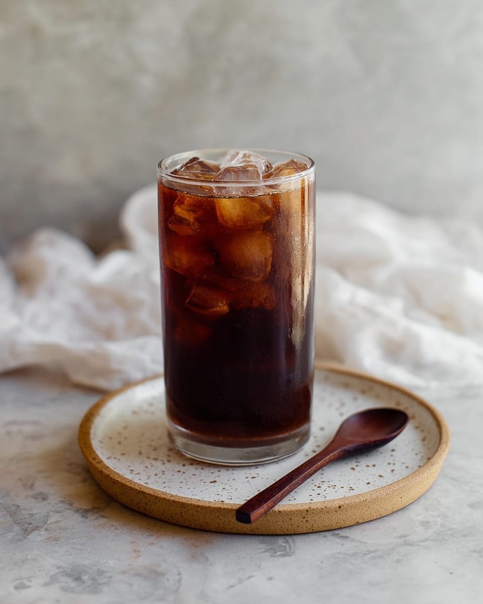 A tall clear glass filled with dark brown iced coffee, showing several ice cubes floating and some submerged inside. The glass sits on a round white plate with a speckled pattern and a thin raised edge. A small wooden spoon with a deep brown color is placed diagonally on the plate in front of the glass. The background shows a soft, blurred white marbled surface with a crumpled white cloth slightly visible behind the glass. The lighting is natural and soft, giving the coffee a cool and refreshing look photo taken with an iphone --ar 4:5 --v 7