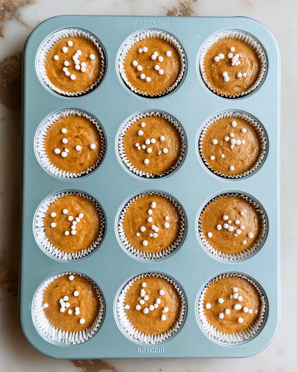 A light blue muffin tray holds twelve unbaked cupcakes, each in a white paper liner. The batter is light brown and smooth, filling each liner almost to the top. On the surface of each cupcake, there are small clusters of white round sprinkles scattered evenly. The tray is placed on a white marbled surface, which has subtle gray and brown veins. The image is clear and bright, showing a top-down view of the cupcakes. photo taken with an iphone --ar 4:5 --v 7