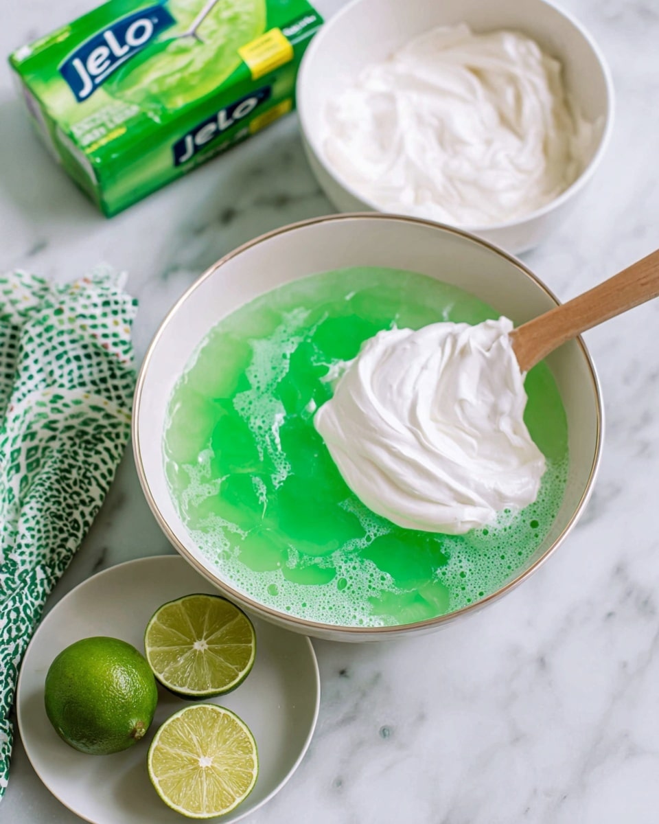 The image shows a mixing bowl with two visible layers, a bright green lime Jello layer at the bottom with bubbles and a smooth, thick white cream layer on top, partially mixed with a wooden spatula. Behind it is a white bowl with more white cream. To the left, there is a bright green box of lime Jello and a folded cloth with a green and white pattern. In front of the bowl, there is a white plate holding one whole lime and two lime halves, showing their juicy, light green inside. The setting is on a white marbled surface photo taken with an iphone --ar 4:5 --v 7