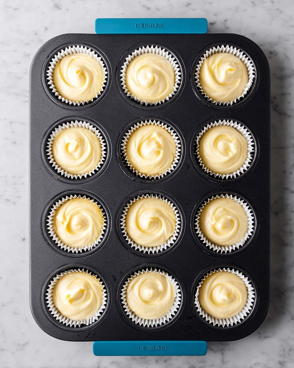 A black muffin tray with twelve slots holds twelve cupcake liners filled with smooth, creamy batter. Each batter portion swirls gently and is pale yellow and creamy white in color, creating soft patterns inside the white paper liners. The tray rests on a white marbled surface. The top and bottom edges of the tray have blue handles with the brand name visible. photo taken with an iphone --ar 4:5 --v 7