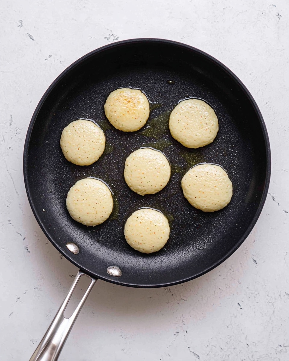 A black frying pan is placed on a white marbled surface with seven small, round batter blobs cooking inside. These blobs are light beige with tiny darker specks and have a smooth, slightly shiny texture. The pan handle is silver metal with a hole near the end. There is a small amount of oil visible around the batter pieces inside the pan. photo taken with an iphone --ar 4:5 --v 7