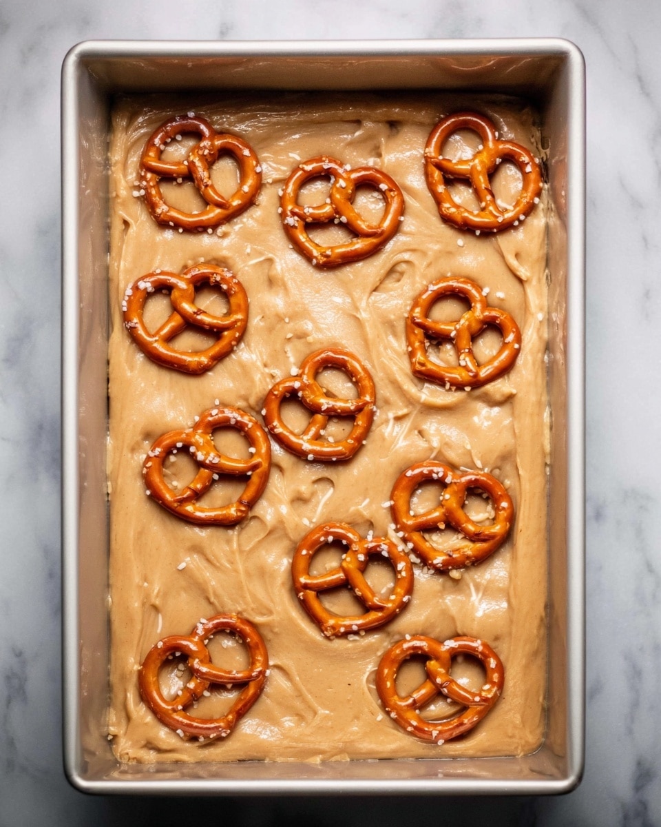 The image shows a rectangular white baking pan filled with a thick, light brown dough layer. On top of the dough, there are several whole pretzels arranged scattered evenly across the surface. The dough has a smooth, slightly shiny texture, and the pretzels are golden brown with a slightly glossy finish. The background is a white marbled surface. Photo taken with an iphone --ar 4:5 --v 7