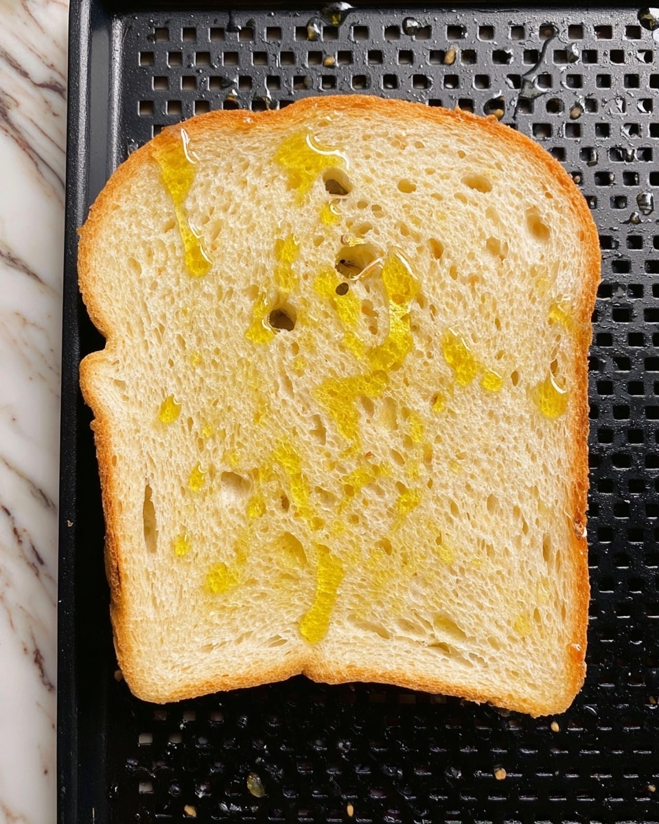 A single slice of pale yellow bread with small air holes scattered throughout is shown laying flat on a black baking tray with grid-like holes. Thin glistening drops of golden oil are drizzled unevenly on the bread surface. The background features a white marbled texture. photo taken with an iphone --ar 4:5 --v 7