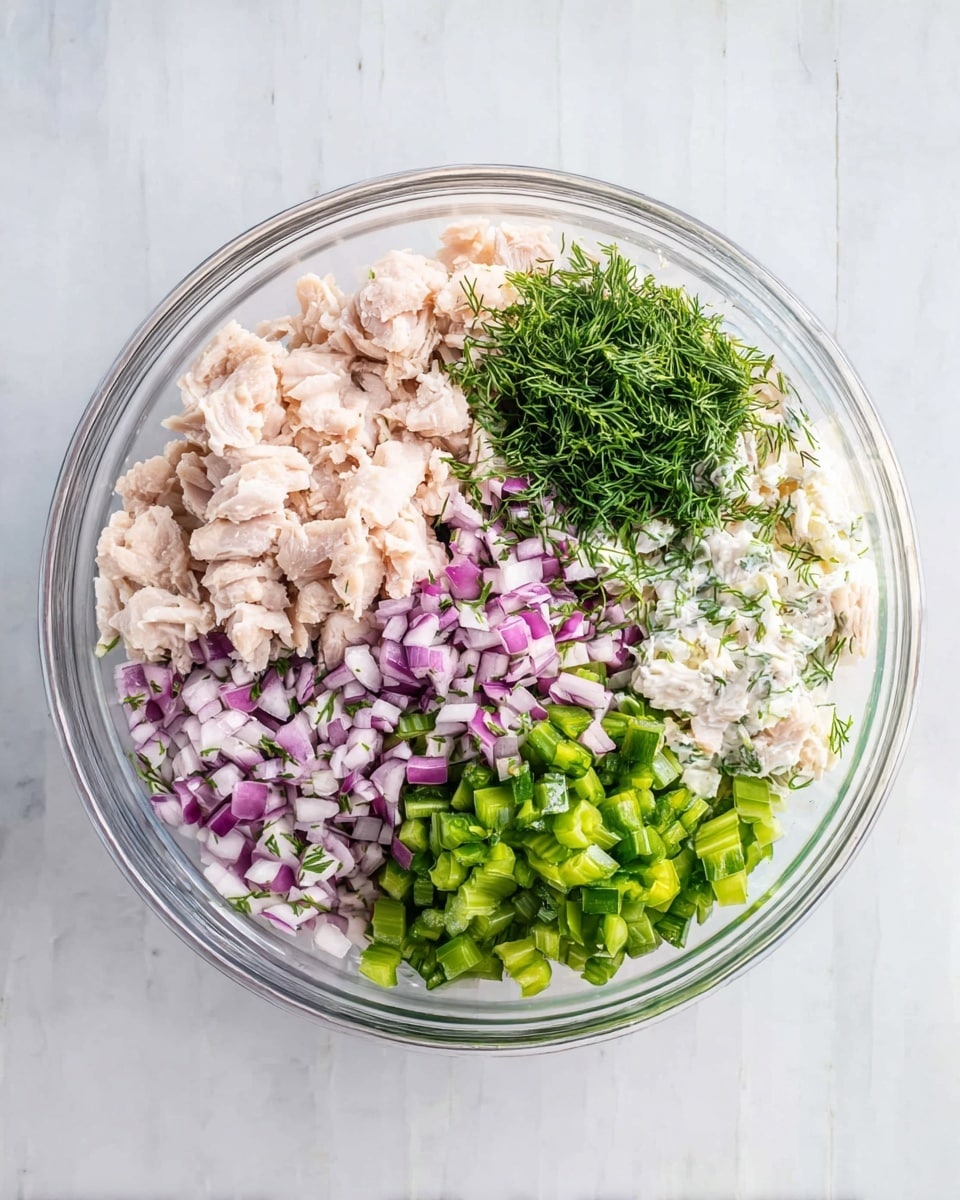 A clear glass bowl sits on a white marbled surface, holding five separate layers of ingredients neatly arranged. On the left side, there are light pink small pieces of chopped cooked chicken. Adjacent to that, bright green chopped celery pieces form a dense layer. To the right of the celery, finely chopped purple-red onion pieces add a colorful contrast. Above the onion, there is a layer of fresh green chopped dill herbs. Near the top edge, a small amount of white mayonnaise or creamy dressing peeks out, adding a smooth texture. Each layer is distinct and fresh, captured in bright natural light. Photo taken with an iphone --ar 4:5 --v 7