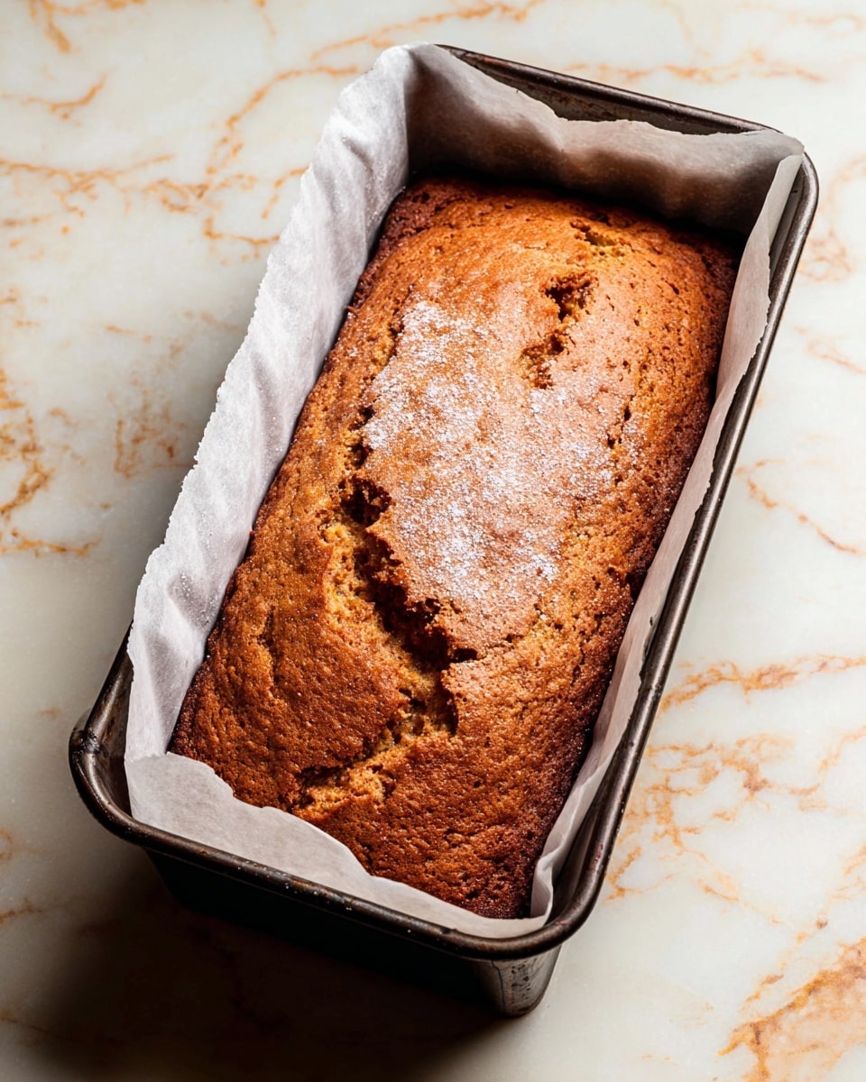 A golden brown loaf cake with a cracked top texture sits inside a dark metal rectangular baking pan lined with white parchment paper that rises above the edges. The cake fills the pan fully, showing a slightly rough, crisp surface with a few light patches of powdered sugar. The pan rests on a white marbled textured surface with faint gray and light orange veining, creating a calm and clean background. photo taken with an iphone --ar 4:5 --v 7