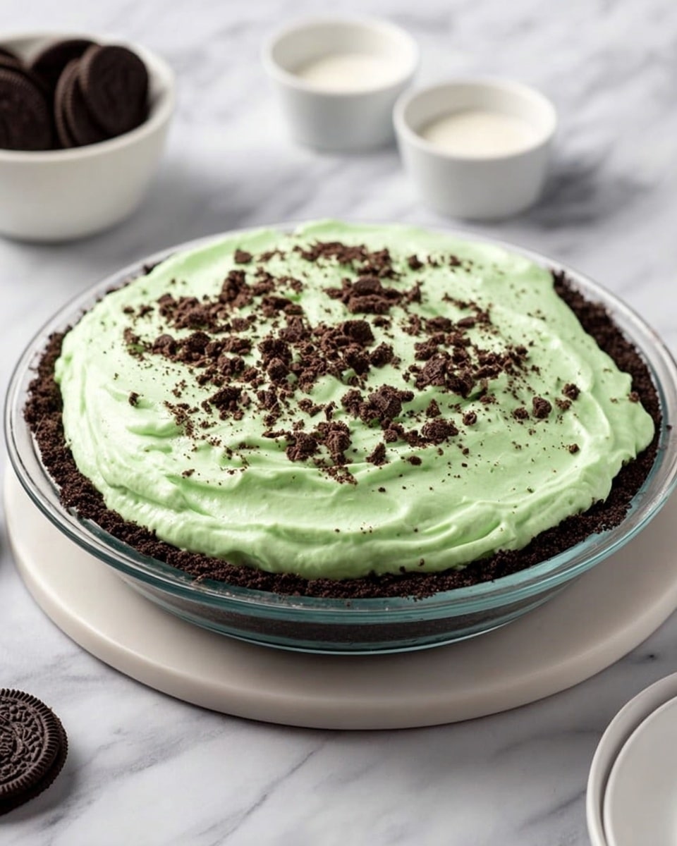 A slice of dessert with three layers sits on a white plate with a white marbled texture background. The bottom layer is a dark crumbly crust, the middle layer is thick, smooth, and light green, and the top has dark chocolate sauce drizzled vertically down the sides. A swirl of white whipped cream with small dark crumbs is placed on top. Some green filling and chocolate sauce have lightly spilled onto the plate. In the background, there is another slice of the same dessert on a stack of white plates and a small white pitcher filled with dark sauce. Photo taken with an iphone --ar 4:5 --v 7