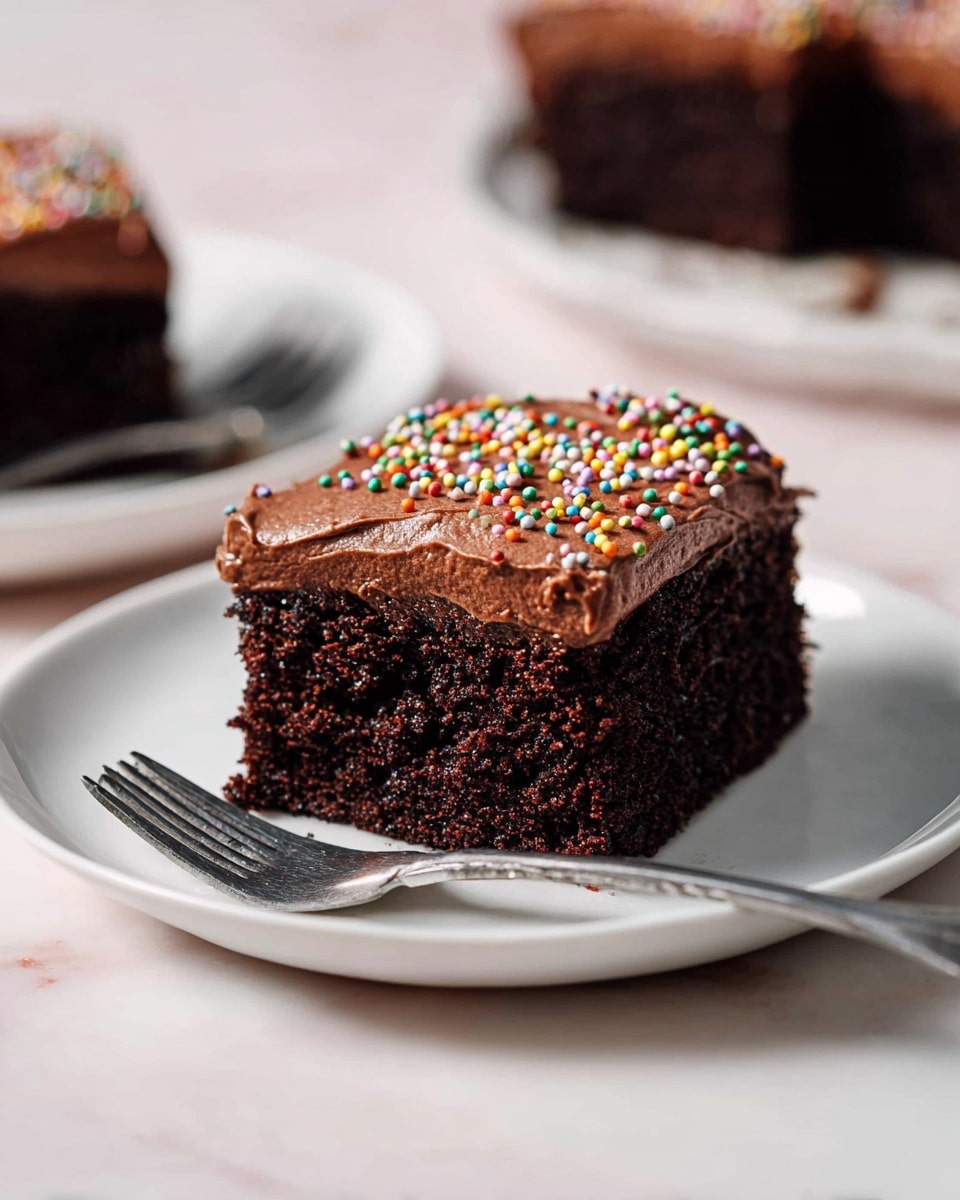 A square piece of dark brown chocolate cake with a thick layer of smooth, creamy chocolate frosting on top. The frosting is decorated with small, round, colorful sprinkles scattered across its surface. The cake looks soft and moist with a slightly crumbly texture. This single piece is placed in the center of a plain white plate on a white marbled surface, accompanied by a silver fork resting beside it. The photo is taken close-up with a soft blurred background showing similar cake pieces on white plates photo taken with an iphone --ar 4:5 --v 7