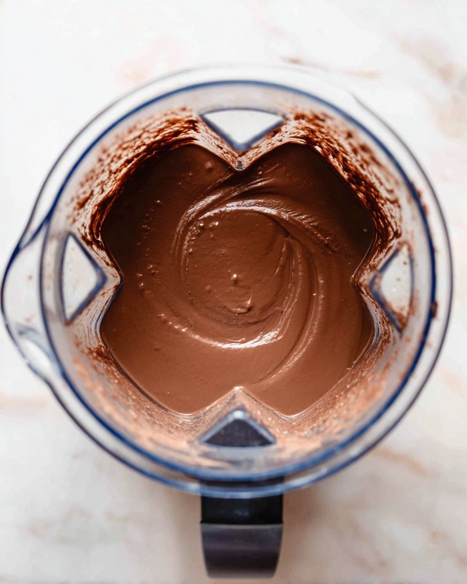 A clear blender container filled with a smooth, shiny chocolate mixture that has a thick and creamy texture, its rich brown color spreading evenly inside. The chocolate fills the container about halfway and has a slight swirl in the center, creating a glossy patch reflecting light. The edges of the container show some thin chocolate streaks where the mixture touched the sides. The blender sits on a white marbled surface, giving a clean and bright background. photo taken with an iphone --ar 4:5 --v 7