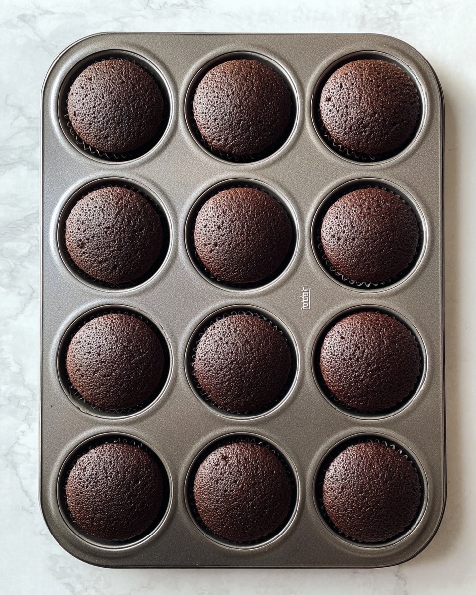 A metal baking tray with twelve round chocolate cupcakes evenly placed in three rows of four, each cupcake having a smooth, slightly domed top with a dark brown color and fine crumb texture, the tray resting on a white marbled surface. photo taken with an iphone --ar 4:5 --v 7
