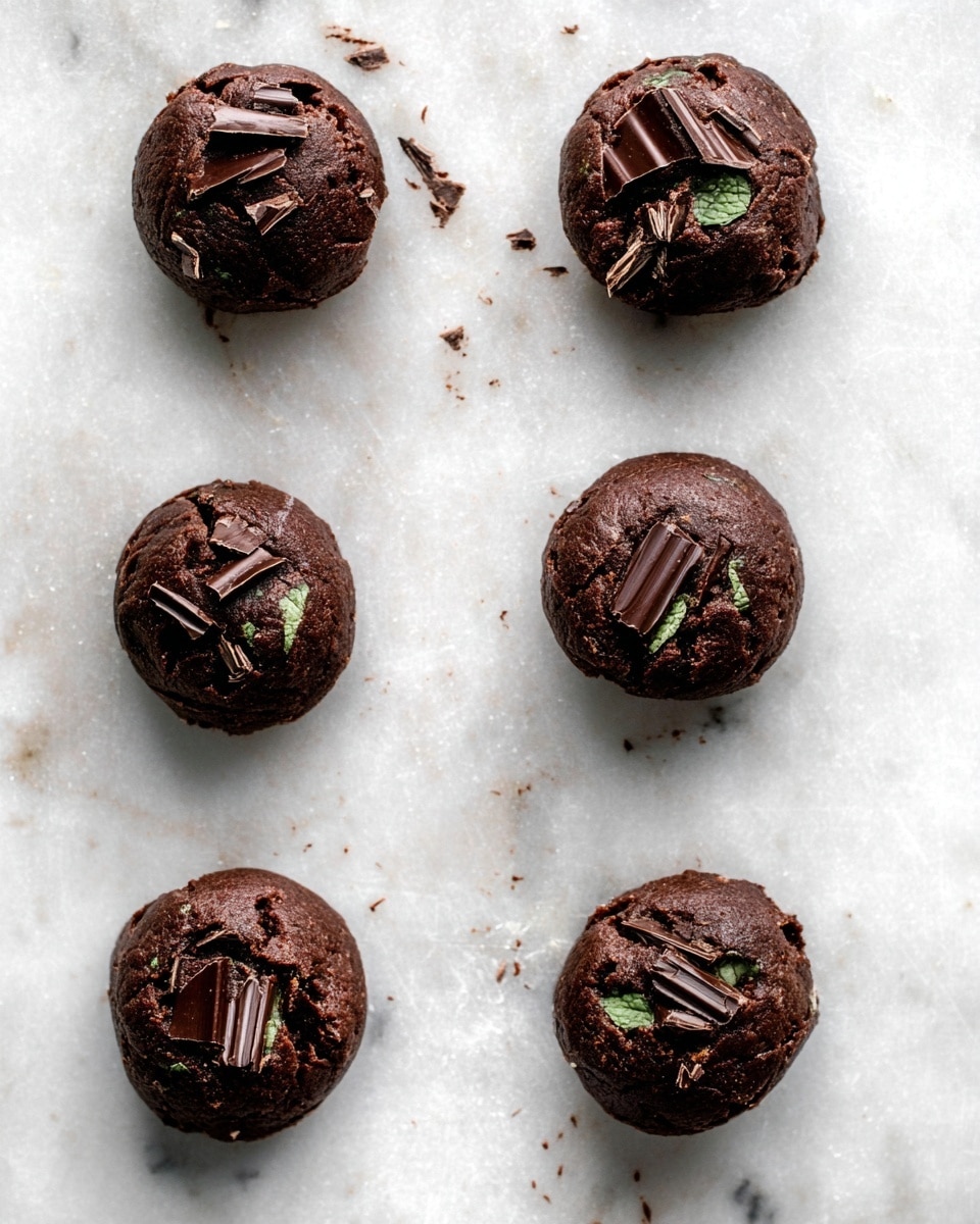 Six round chocolate cookie dough balls sit spaced out on a white marbled surface. Each dough ball is dark brown, thick, and rich in texture, with several pieces of broken chocolate chunks embedded on top, showing layers of dark chocolate and a hint of green mint filling inside some pieces. The dough balls have a slightly uneven and soft surface, appearing thick and moist. The image shows a top-down view with no other items around the cookies photo taken with an iphone --ar 4:5 --v 7
