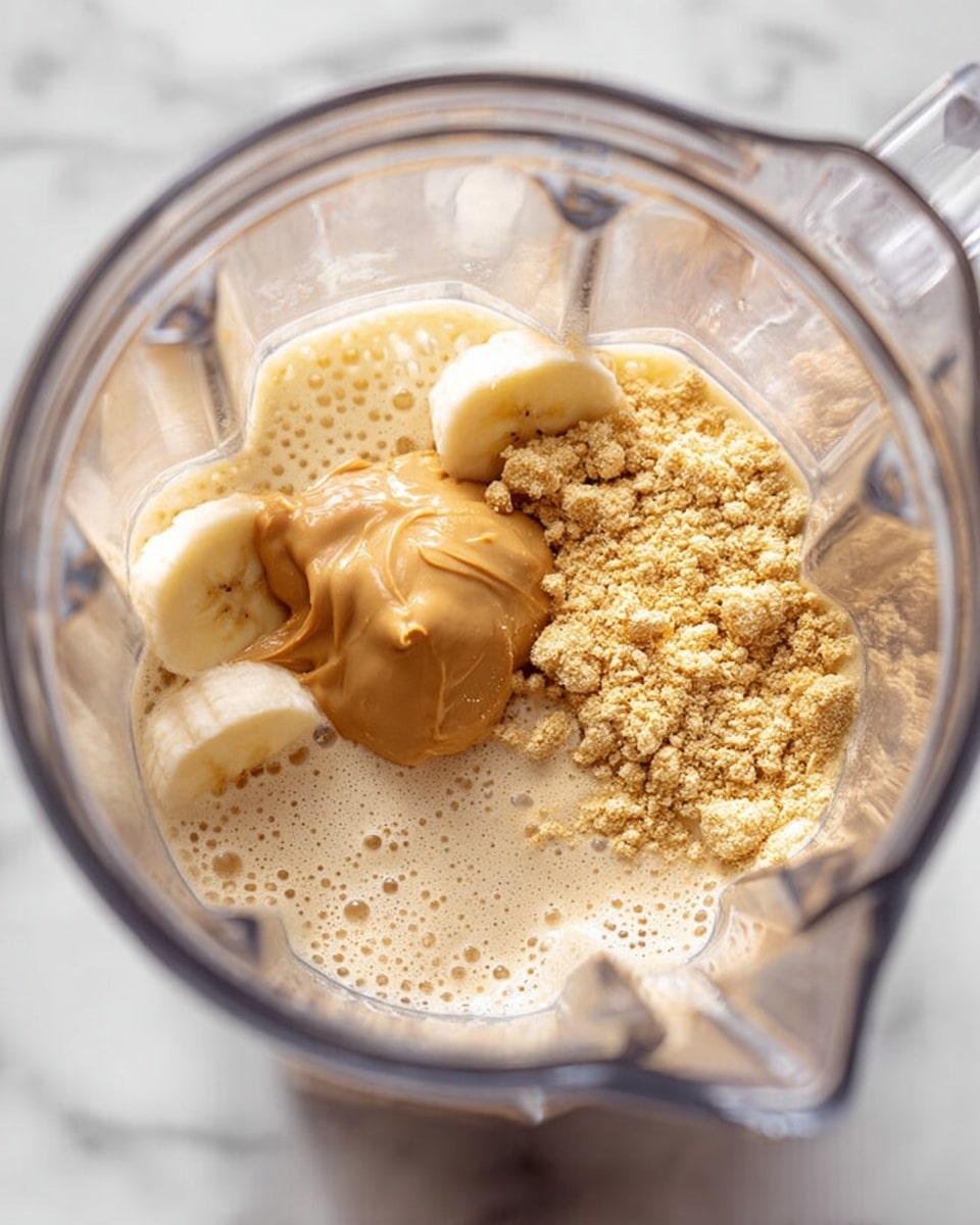 A clear blender container shows several ingredients inside, including a smooth light brown scoop of peanut butter near the bottom left, a whole raw garlic bulb with a light yellowish skin at the top left, a creamy beige powder scattered mostly on the right side, and a foamy layer of light beige liquid filling the rest of the container. The blender is placed on a white marbled surface. photo taken with an iphone --ar 4:5 --v 7