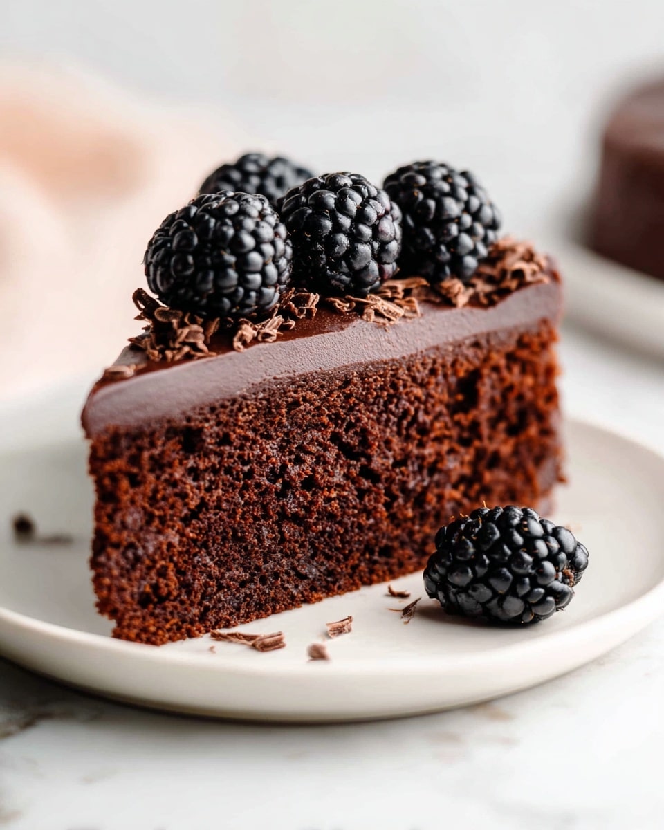 A single slice of dark brown chocolate cake sits on a white plate over a white marbled surface. The cake has one thick layer that looks moist and soft. On top, there is a smooth, glossy layer of dark chocolate frosting that covers the entire top surface of the slice. Three large, shiny blackberries with a bumpy texture are neatly placed in a row on the frosting, with small chocolate shavings scattered around them. One additional blackberry rests beside the cake slice on the plate. The background is light and softly out of focus, enhancing the cake's rich look. Photo taken with an iphone --ar 4:5 --v 7