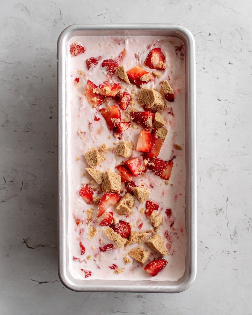 A rectangular white container filled with one thick layer of pink creamy mixture that shows visible pieces of red strawberries throughout. On top of the mixture, there are scattered small chunks of light brown crushed cookies and more chopped strawberry pieces. The container is placed on a white marbled surface. photo taken with an iphone --ar 4:5 --v 7