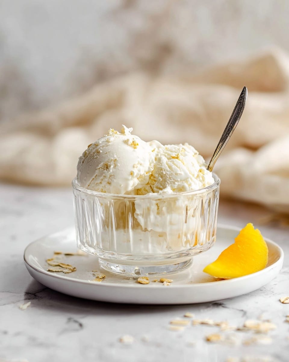 A clear glass bowl with vertical ridges holds two scoops of white ice cream with small light brown bits mixed in. The bowl sits on a white plate scattered with small oat flakes. To the right, a small piece of bright yellow fruit rests on the plate. A silver spoon stands in the ice cream, and the entire setup is on a white marbled surface with a soft beige cloth blurred in the background. Photo taken with an iphone --ar 4:5 --v 7