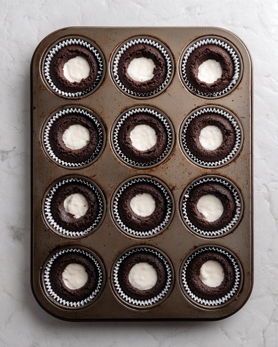 A dark brown chocolate cupcake tray holds twelve cupcakes, each in white paper liners. The tops of the cupcakes have been hollowed out in the center, revealing a smooth, creamy white filling inside the middle of each cupcake. The tray is metal and shows light signs of use. The background is a white marbled surface. photo taken with an iphone --ar 4:5 --v 7
