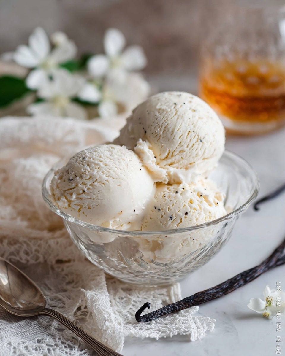 The image shows a clear glass bowl filled with three scoops of creamy, white vanilla ice cream with small black vanilla bean specks visible on the surface. The scoops are slightly rough with a soft texture, stacked closely inside the bowl. To the left of the bowl, there is a silver spoon resting on a white cloth with lace edges. In the background, white flowers and a vanilla pod are visible, placed on a soft beige fabric, and a glass with an amber liquid is blurred behind. The whole setting is on a white marbled texture. photo taken with an iphone --ar 4:5 --v 7