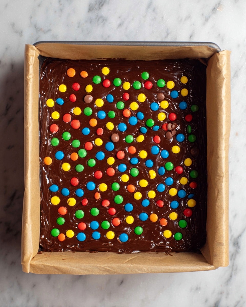 A square baking pan lined with brown parchment paper holds a thick, glossy dark chocolate batter spread evenly inside. The batter is topped with a colorful layer of small candy-coated chocolate pieces scattered all over in red, yellow, green, blue, and orange. The pan rests on a white marbled surface. photo taken with an iphone --ar 4:5 --v 7