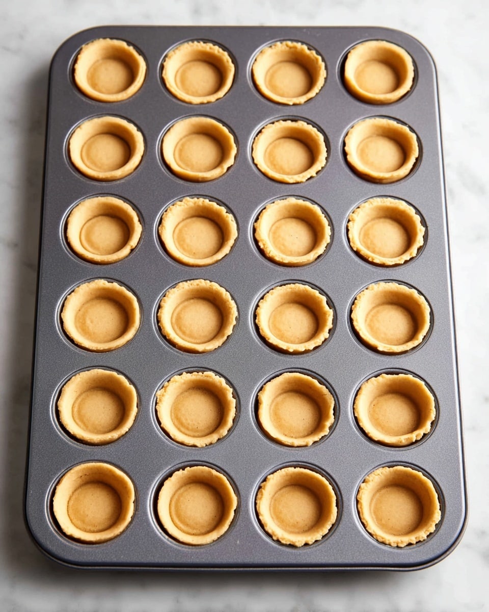 The image shows a grey mini muffin pan filled with 24 small tart shells evenly arranged in four rows and six columns. Each tart shell has a smooth, light golden-brown color with a slightly raised dome in the center, and visible edges pressed neatly against the pan's round molds. The pan is placed on a white marbled surface, and the overall scene is bright with soft lighting. photo taken with an iphone --ar 4:5 --v 7