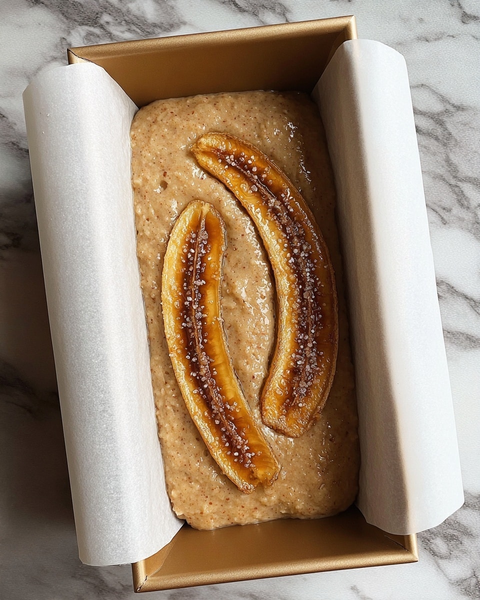 The image shows a loaf pan lined with white parchment paper filled with light brown banana bread batter. On top of the batter are two halved bananas, placed curved side up, with a golden brown caramelized surface, sprinkled with coarse sugar crystals. The pan is set on a white marbled surface. photo taken with an iphone --ar 4:5 --v 7