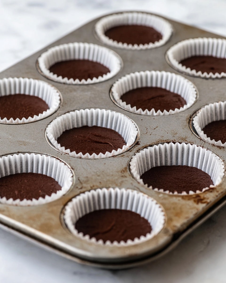 The image shows a metal baking tray with eight round cups lined with white paper liners, each filled with dark brown chocolate batter. The batter is smooth and fills each liner about halfway, creating a flat surface on top. The tray has some light stains, giving it a used look, and it rests on a white marbled surface. The focus is sharpest on the front cups, with the others softly blurred in the background, creating a cozy baking atmosphere. photo taken with an iphone --ar 4:5 --v 7