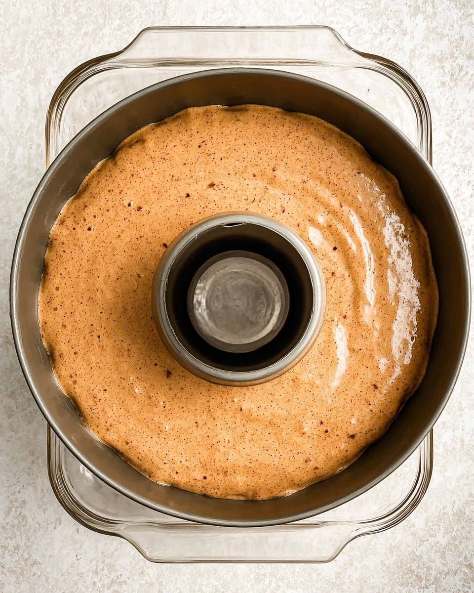 A close-up view of a round, brown batter with a smooth and slightly speckled texture filling a fluted bundt cake pan with a central tube, resting inside a clear rectangular glass baking dish. The batter is evenly spread, showing small dark flecks scattered across its surface. The scene is set on a white marbled texture background. Photo taken with an iphone --ar 4:5 --v 7