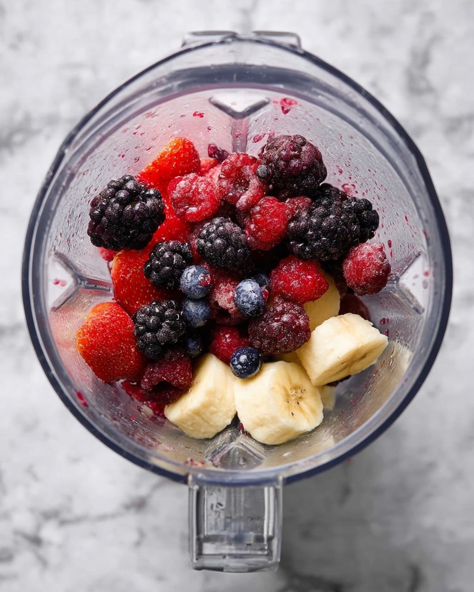 A clear blender jar viewed from above shows two layers of fruit inside: the bottom layer has a few small pale yellow banana pieces, and the top layer is filled with a mix of dark purple blackberries, bright red strawberries, dark red raspberries, and dark blue blueberries. The blender is sitting on a white marbled surface. photo taken with an iphone --ar 4:5 --v 7