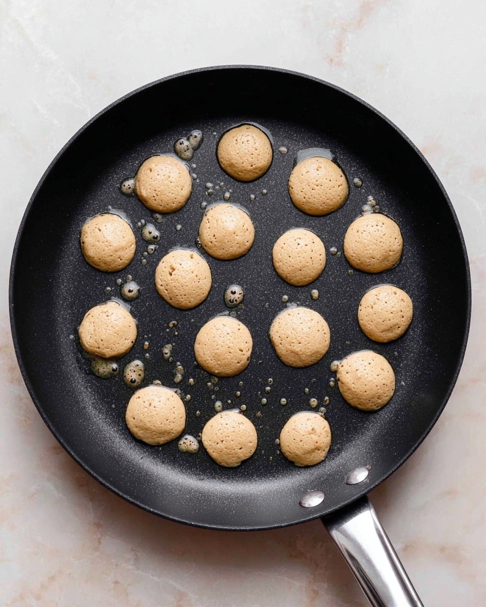 A black non-stick pan with a silver handle is shown on a white marbled surface. Inside the pan, there are sixteen small round pancake-like batter blobs spread out evenly. Each blob is light brown with tiny specks, showing a soft, slightly bubbly texture on top as if they are cooking. Small droplets of oil are visible around the batter pieces, giving the pan a slightly shiny look. Photo taken with an iphone --ar 4:5 --v 7