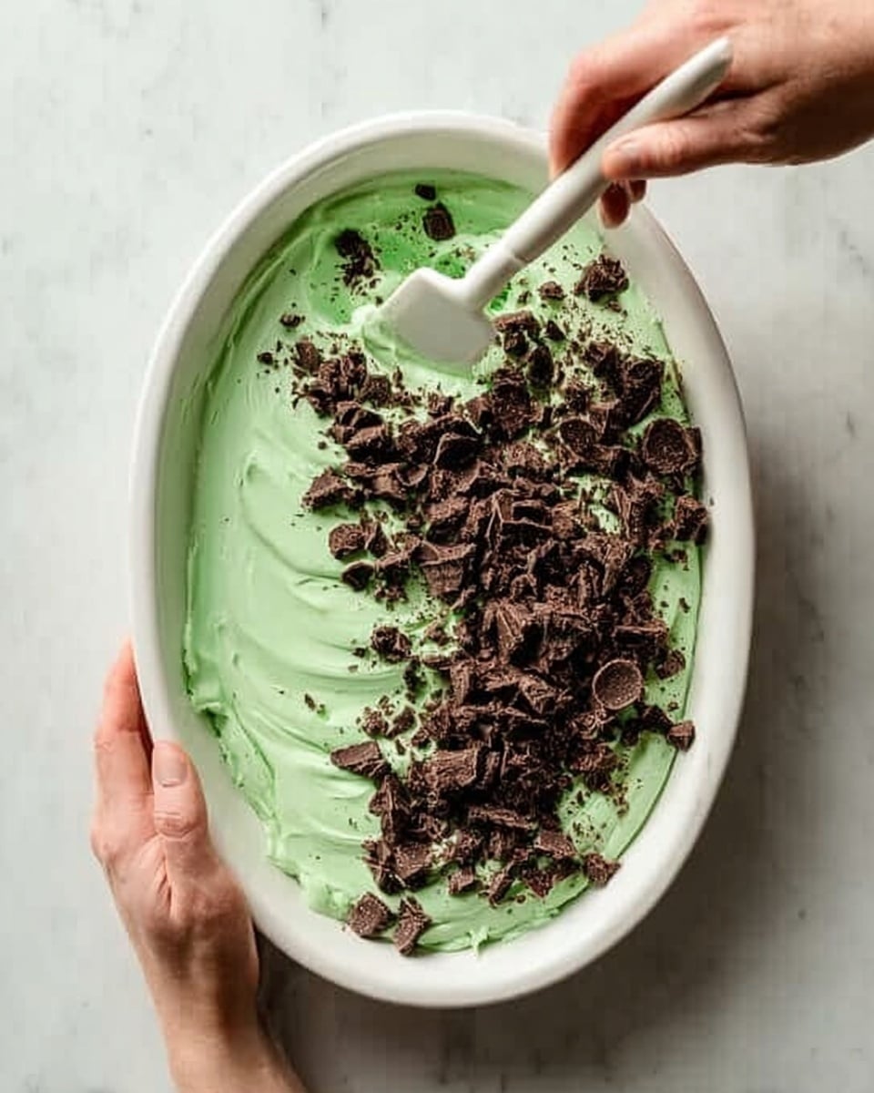 A white oval dish filled with one thick layer of light green creamy mixture. On top, there is a pile of chopped dark brown chunks spread mostly on one side of the green creamy layer. A woman's hand is holding the dish steady on the left side, while the other woman’s hand is using a white spatula to mix or spread the green mixture near the dark chunks. The background is a white marbled surface. photo taken with an iphone --ar 4:5 --v 7