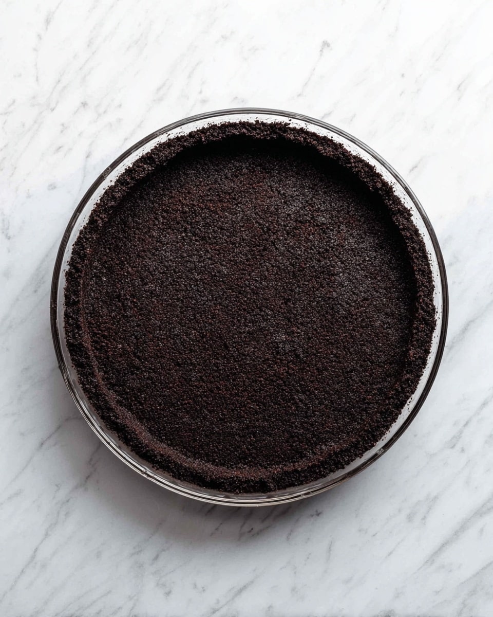 A single dark brown layer of crushed cookie crust is pressed evenly against the bottom and sides of a clear round pie dish, creating a firm and textured base for a dessert. The pie dish is placed on a white marbled surface, and the dark crust contrasts sharply with the white background. Photo taken with an iphone --ar 4:5 --v 7