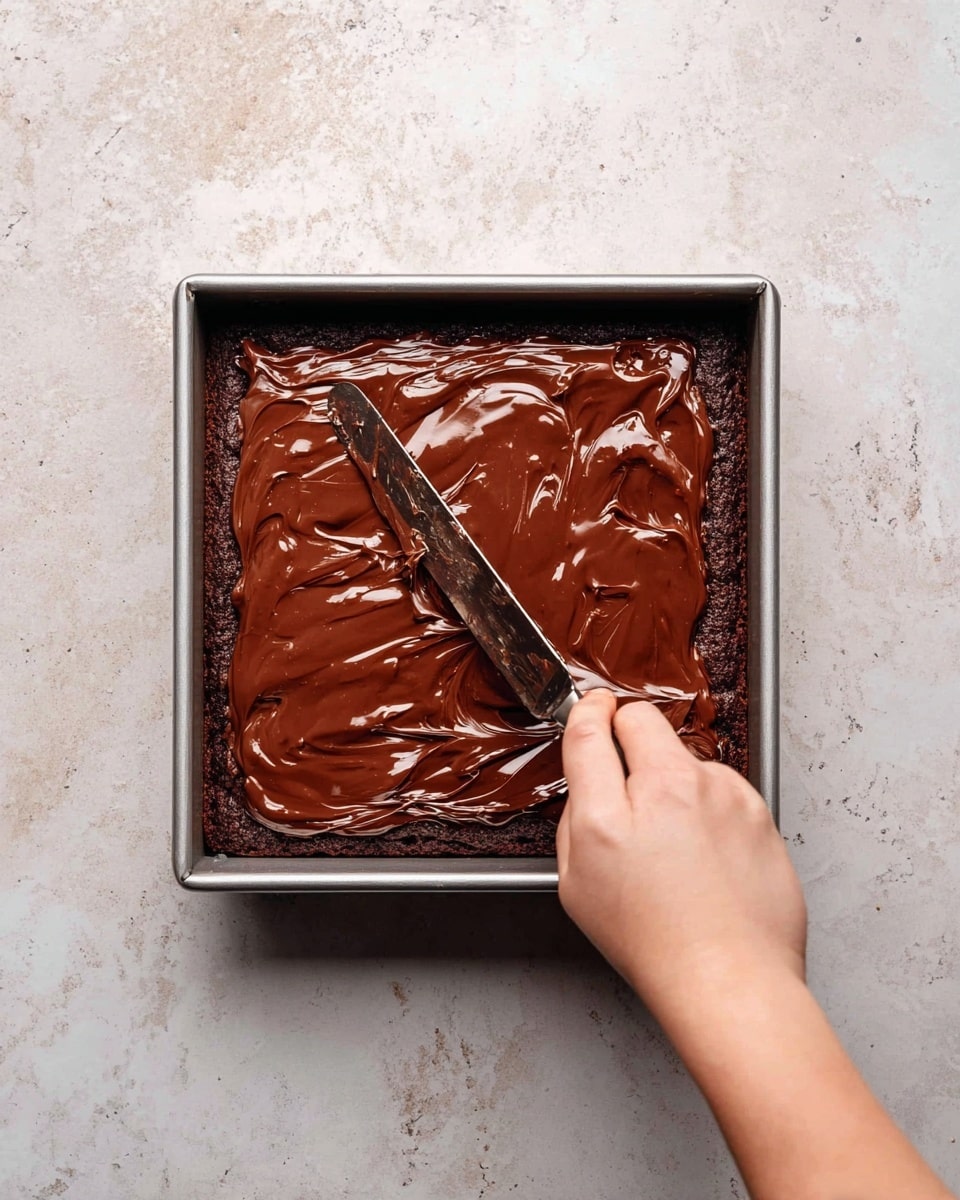 A square metal pan holds a thick, dark brown chocolate cake base as the bottom layer, with smooth, glossy, rich chocolate frosting being spread evenly on top with a knife by a woman's hand; the chocolate frosting layer is shiny, thick, and slightly uneven with some swirls and texture. The pan sits on a white marbled surface. photo taken with an iphone --ar 4:5 --v 7