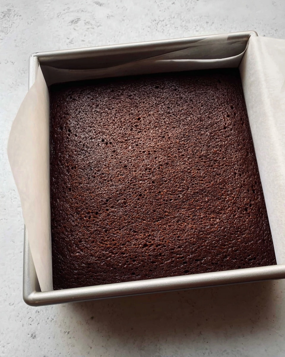 A freshly baked dark brown chocolate cake sits inside a square metal baking pan lined with white parchment paper that is slightly folded back on one corner, revealing the cake's spongy texture with small air holes on the surface. The pan rests on a white marbled surface, highlighting the rich, moist-looking texture of the cake that fills the entire pan with even thickness and smooth edges, no decoration or frosting yet. Photo taken with an iphone --ar 4:5 --v 7