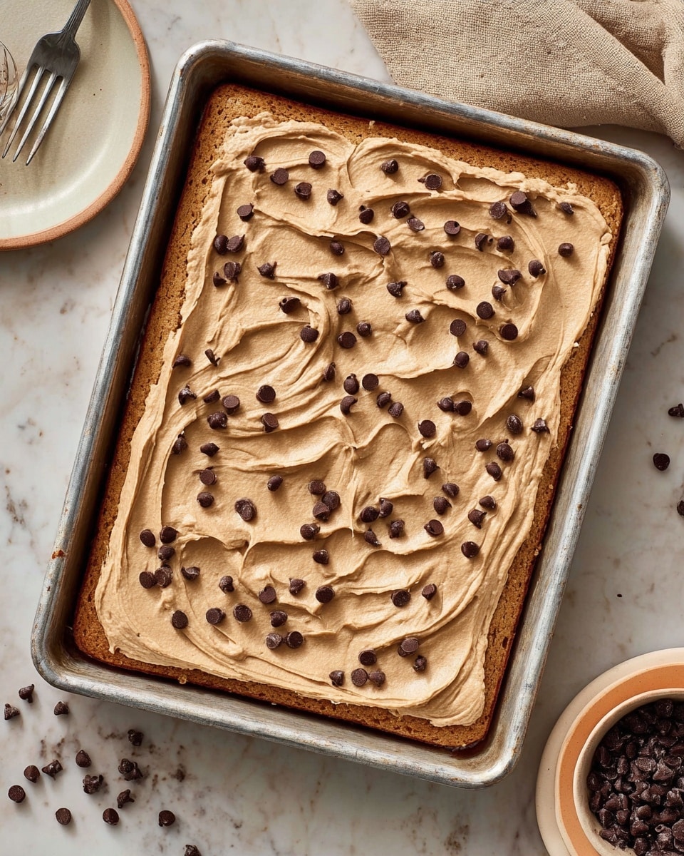 A rectangular metal baking tray holds a baked bar with two visible layers; the bottom layer is golden brown and looks soft and thick, while the top layer is a light tan frosting spread smoothly with swirls, sprinkled with small dark chocolate chips evenly on top. The tray sits on a white marbled surface with a beige plate holding a spoonful of dark chocolate chips to the side, and part of a woman's hand holding a fork is visible in the top left corner. photo taken with an iphone --ar 4:5 --v 7