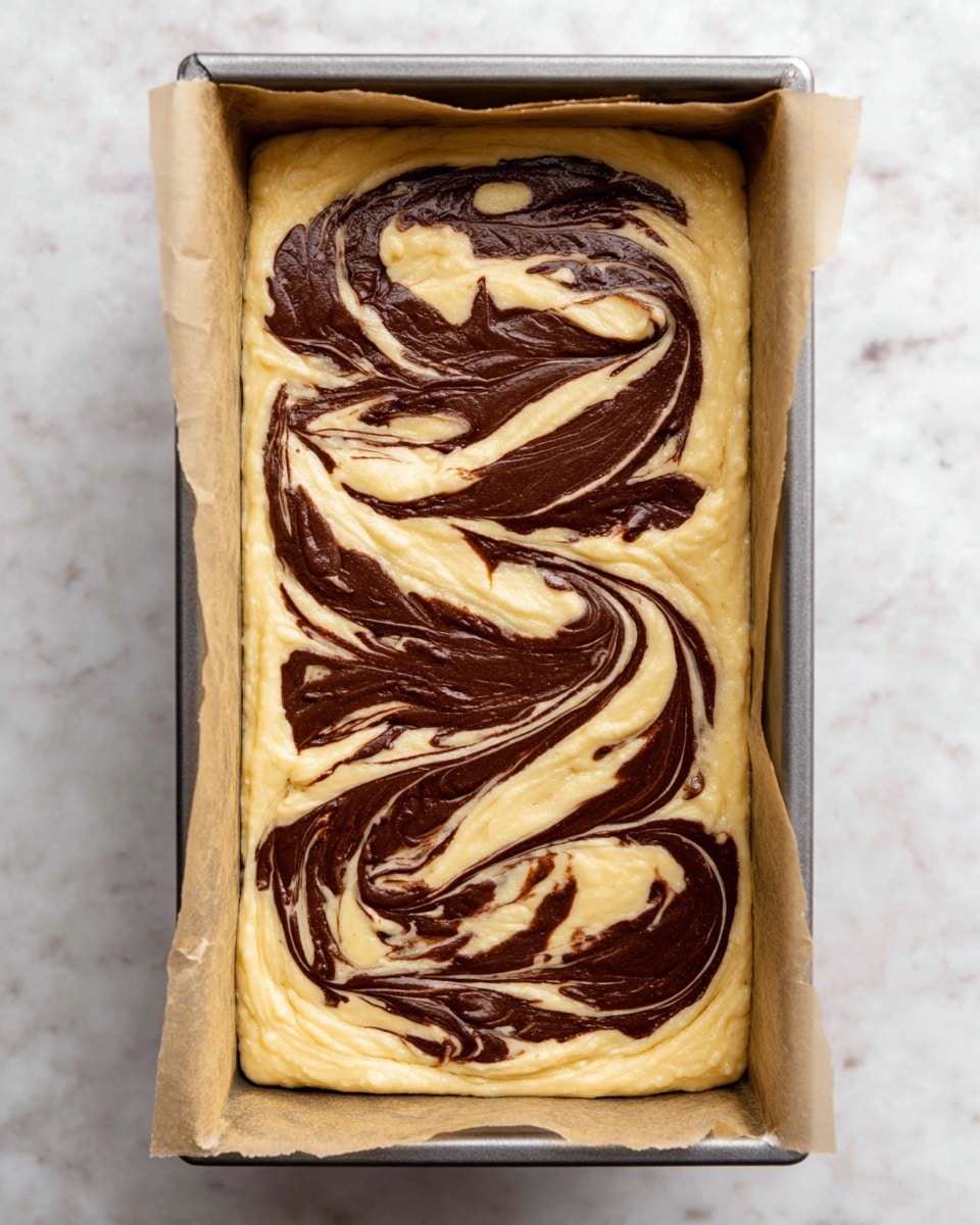 The image shows a rectangular metal baking pan lined with parchment paper, filled with two layers of batter. The base layer is light yellow with a smooth texture, while the top layer is a dark brown chocolate swirl spread in a flowing, artistic pattern. The swirls contrast sharply with the pale yellow base, creating a marbled effect across the entire surface. The pan sits on a white marbled surface. Photo taken with an iphone --ar 4:5 --v 7