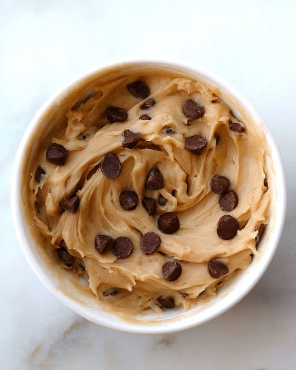 The image shows a white bowl filled with a smooth, creamy light brown dough mixture that has several dark brown chocolate chips mixed and scattered on top. The dough has soft swirls and folds visible, with the chocolate chips sitting prominently on the surface. The bowl is placed on a white marbled surface. photo taken with an iphone --ar 4:5 --v 7