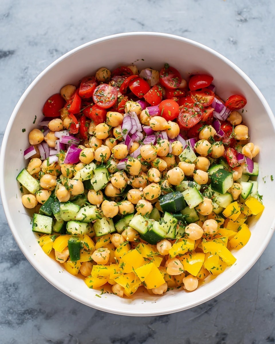 A white bowl filled with a fresh salad showing four main layers: the base layer has round, beige chickpeas, the middle layers are brightly colored with chopped yellow bell peppers and green cucumber pieces, and the top layer has small, halved red cherry tomatoes and finely chopped red onions scattered around. Small green herb bits are sprinkled evenly throughout. The bowl sits on a white marbled surface. Photo taken with an iphone --ar 4:5 --v 7