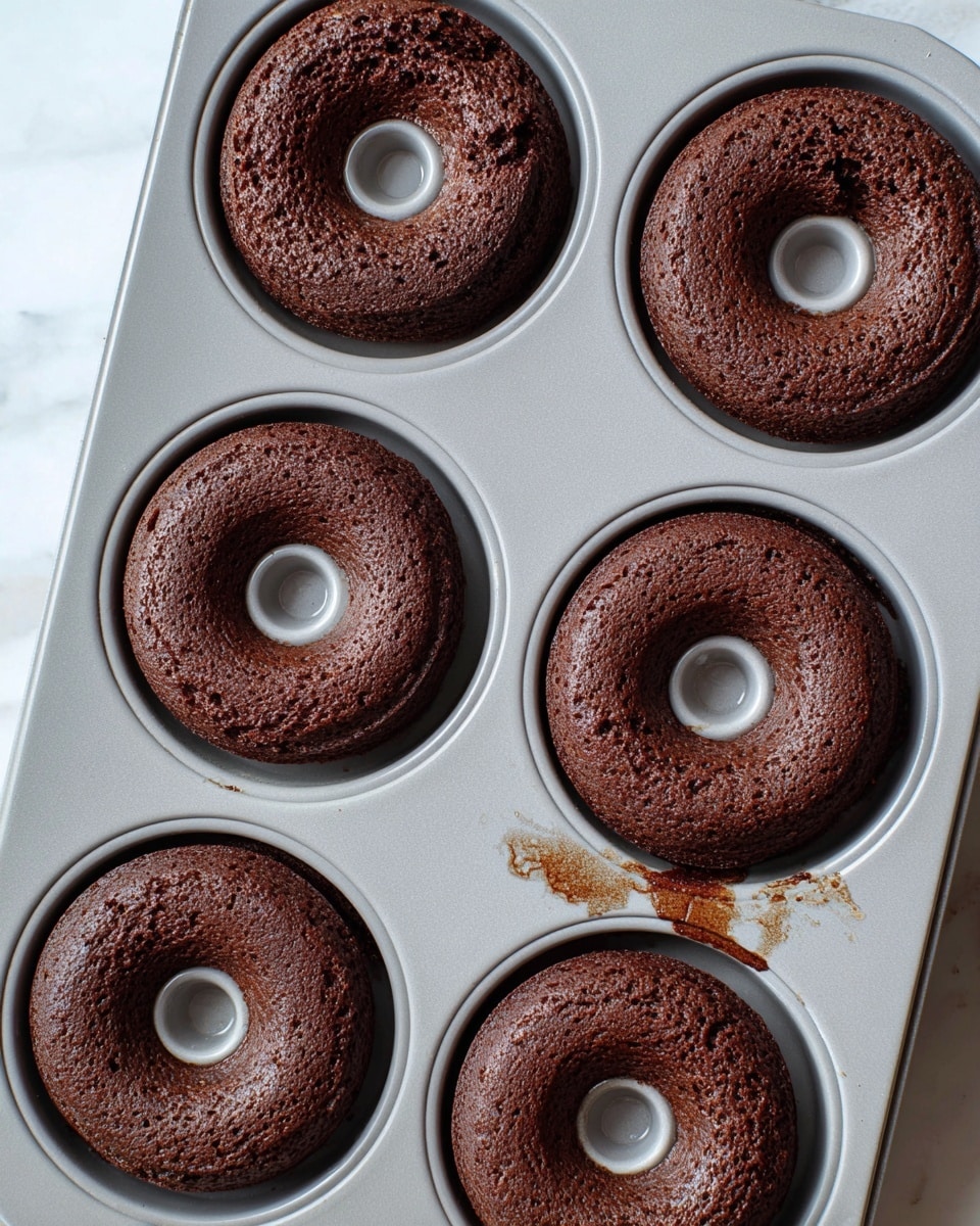 The image shows four freshly baked chocolate cake donuts in a light gray silicone donut pan placed on a baking tray. Each donut is rich dark brown with a slightly cracked and soft texture on top, and each has a smooth raised center hole from the donut mold. The pan rests on a white marbled surface which contrasts with the dark brown color of the donuts. The donuts' edges touch the round mold, showing slight unevenness in the baking. There is some moisture or droplets visible on the pan surface near the donuts. photo taken with an iphone --ar 4:5 --v 7