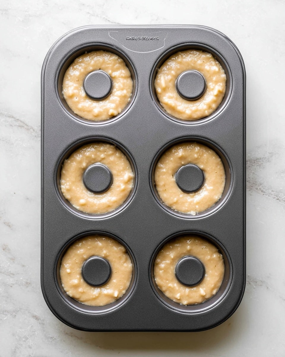 A dark grey metal donut baking tray with six donut cavities is filled with a light brown batter with a slightly bubbly texture, each cavity almost full with batter. The tray sits on a white marbled surface, and the batter has a creamy, smooth look with small lumps visible. The centers have a raised darker grey cylindrical piece. There are no extra objects or hands in the image. photo taken with an iphone --ar 4:5 --v 7