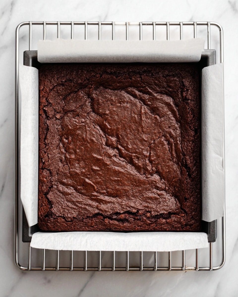 A square chocolate brownie with a rich, dark brown color and a slightly cracked and wrinkled top layer, sitting inside a white parchment paper-lined square baking pan placed on a metal cooling rack, all set on a white marbled surface. The brownie looks dense and moist with an even, thick texture covering the whole pan. Photo taken with an iphone --ar 4:5 --v 7