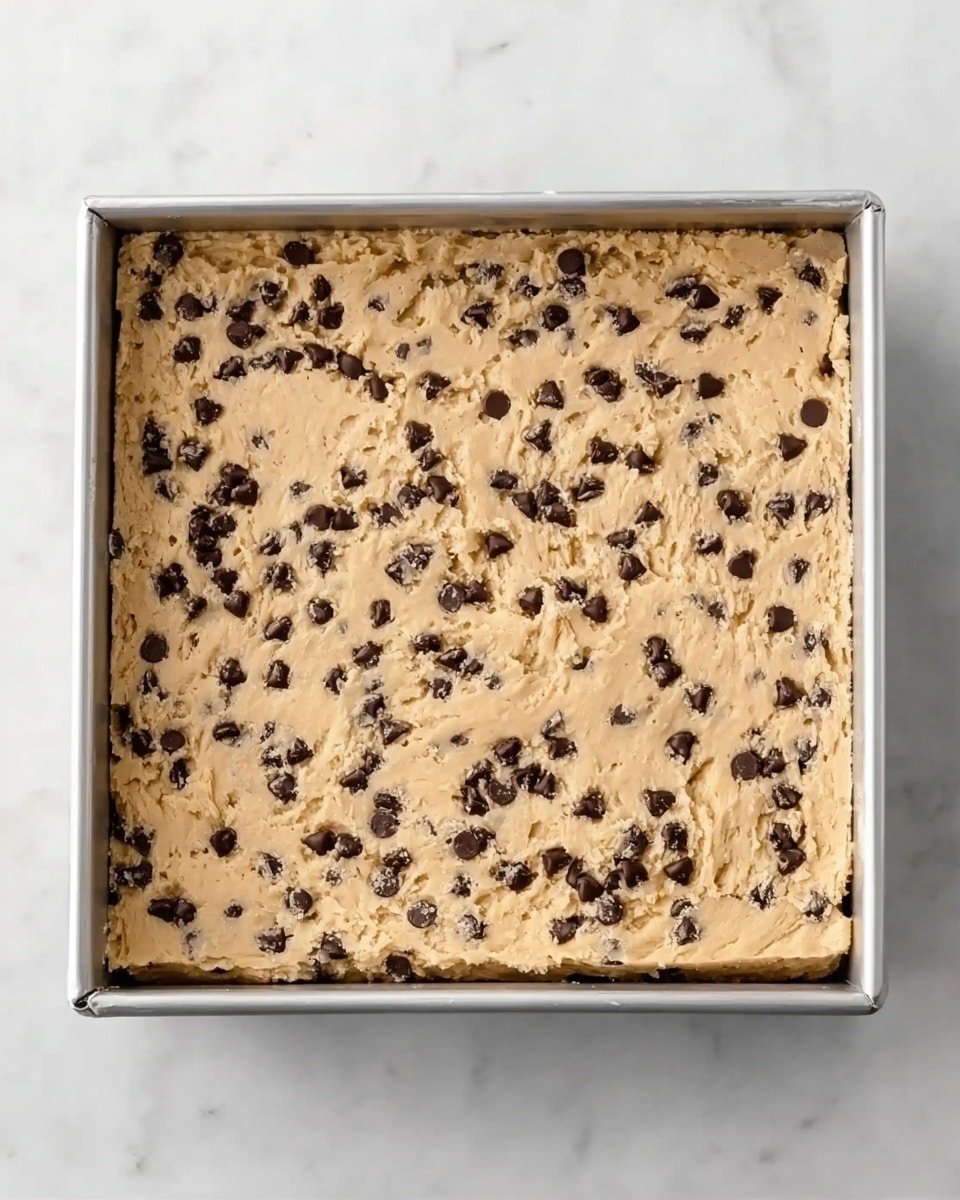 The image shows a square metal baking pan filled with an even layer of raw cookie dough that has small chocolate chips spread throughout. The dough has a light tan color with dark chocolate chips creating a speckled pattern. The pan is placed on a white marbled surface, giving a clean and bright background. The dough reaches the edges of the pan, making a smooth, flat top layer. photo taken with an iphone --ar 4:5 --v 7