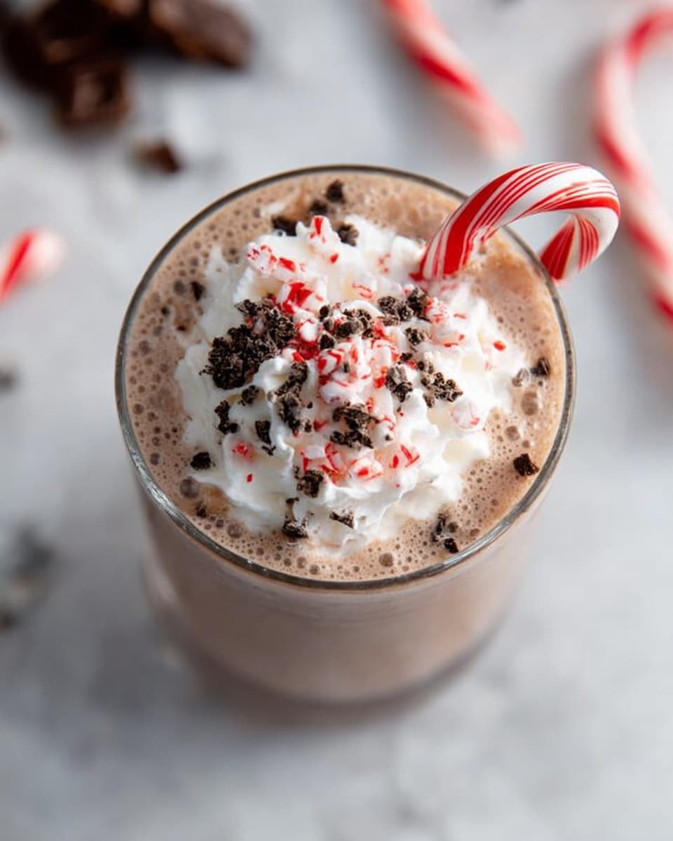 A close-up view of a creamy chocolate drink in a clear glass, filled almost to the top with a light brown, smooth liquid. The drink is topped with a generous layer of white whipped cream that has soft, fluffy peaks. On top of the whipped cream, there are small pieces of crushed dark chocolate and tiny red and white candy cane bits scattered evenly. A red and white striped candy cane is placed inside the glass, leaning slightly to the side. The glass is set on a white marbled surface, with a blurred background showing broken pieces of chocolate and another candy cane. photo taken with an iphone --ar 4:5 --v 7